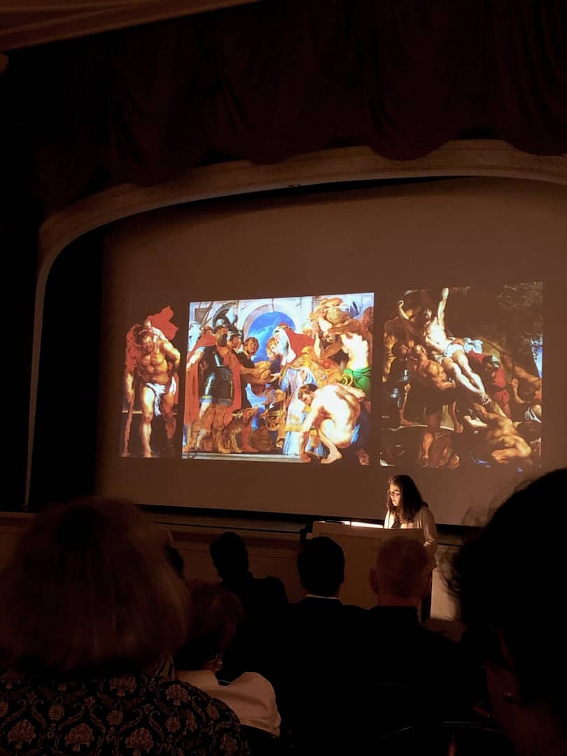 A speaker stands at a lecturn in front of a screen filled with projected images of paintings by Rubens.
